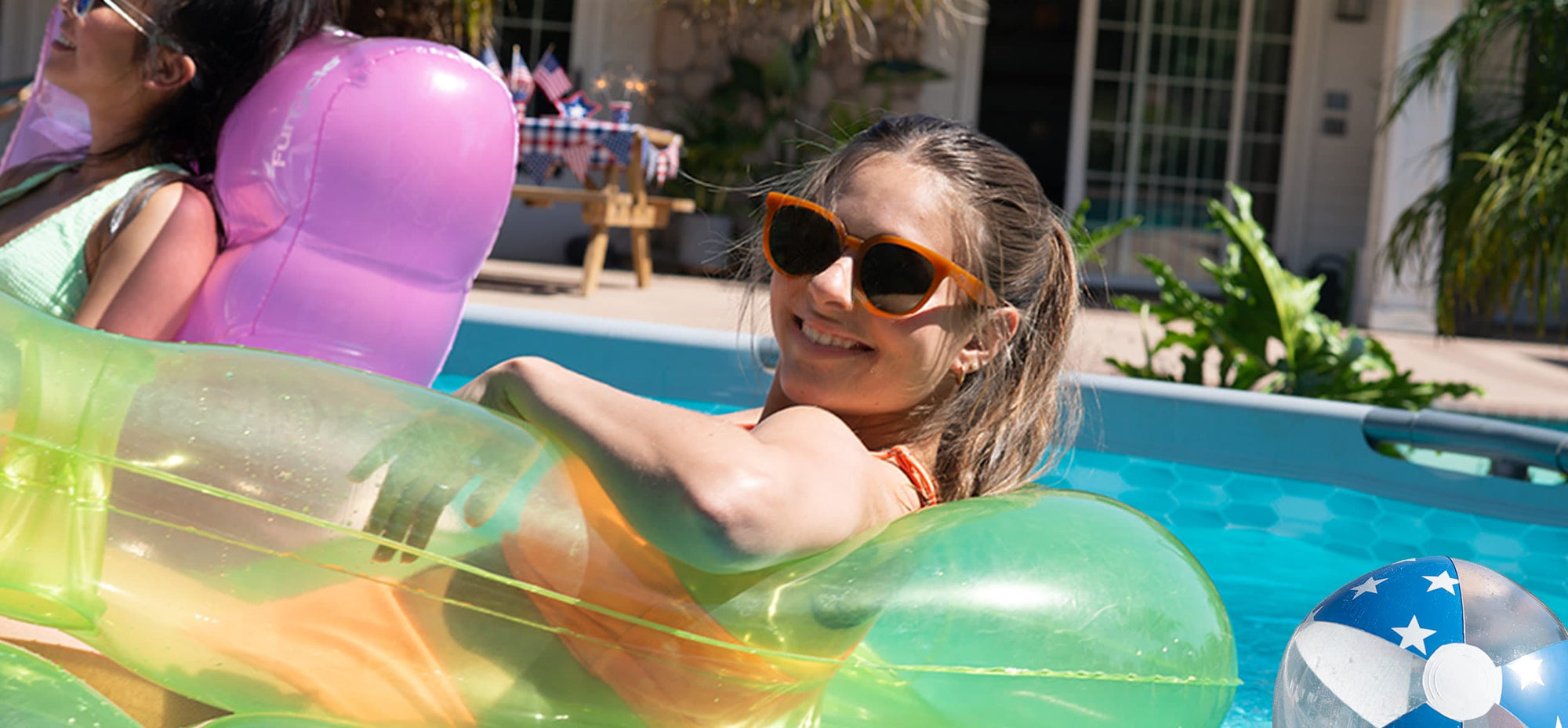 a woman smiling to the camera while lounging in an inflatable inside Funsicle above ground pool with 4th July picnic table in the background