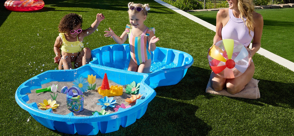 two kids playing in a sand pool filled with paper flowers with mom watching them from the side