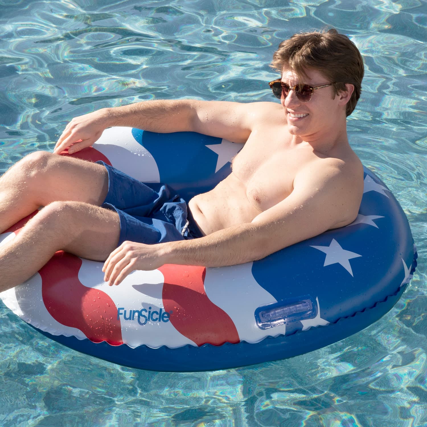 Man in sunglasses and blue shorts sitting on a red, white, Funsicle inflatable tube in clear water.